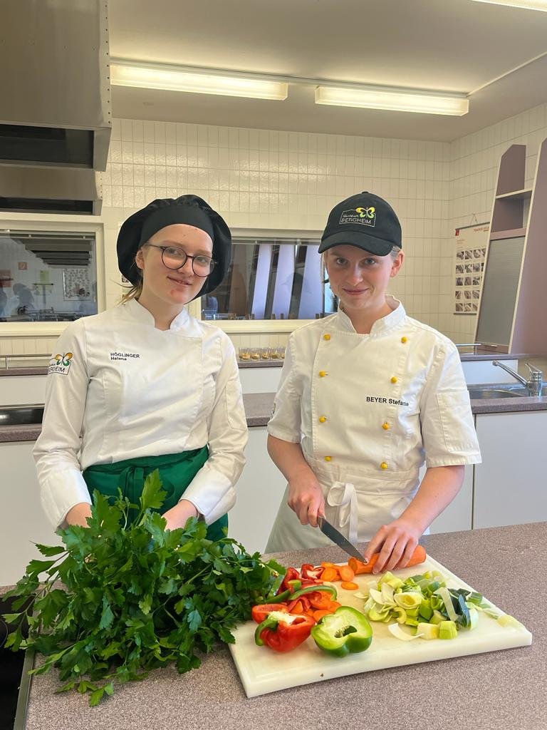 zwei Mädchen in Kochkleidung schneiden buntes Gemüse und Kräuter